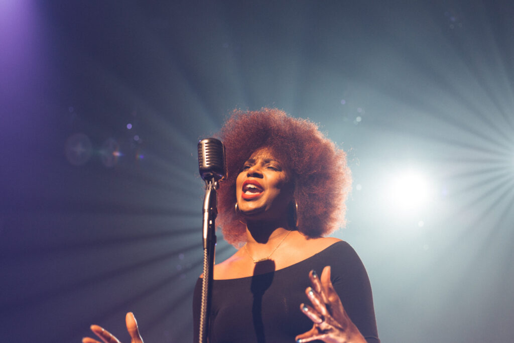 black vocalist singing into a microphone with stage lights behind her